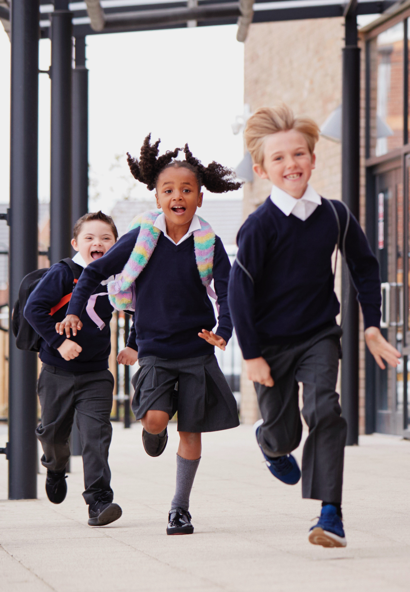 Primary school kids, wearing school uniforms and backpacks, running on a walkway outside their school building, front view